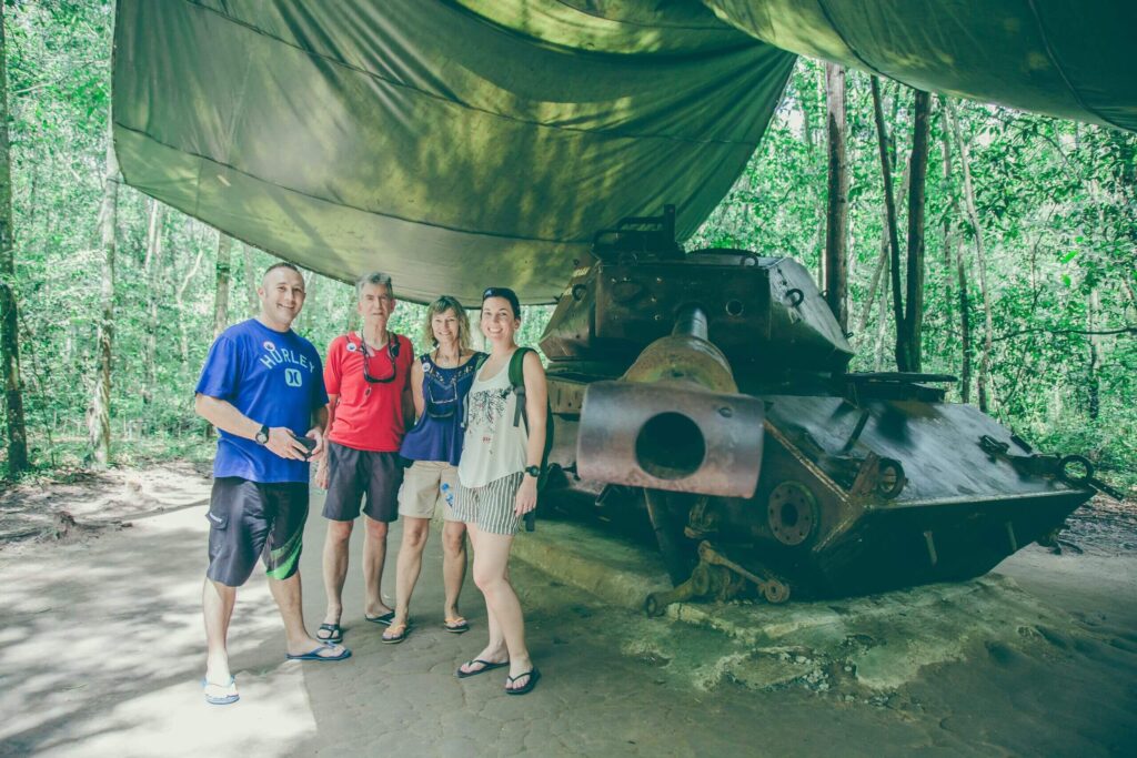A group of four tourists posing in front of a rusted, historical Vietnam War tank under a shaded forest canopy during a guided Cu Chi Tunnels tour from Ho Chi Minh City with Saigon Adventure.