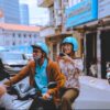 A smiling tourist taking photos while riding on the back of a motorbike with a local student guide during a scooter tour in Ho Chi Minh City.
