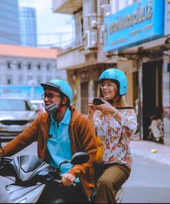 A smiling tourist taking photos while riding on the back of a motorbike with a local student guide during a scooter tour in Ho Chi Minh City.