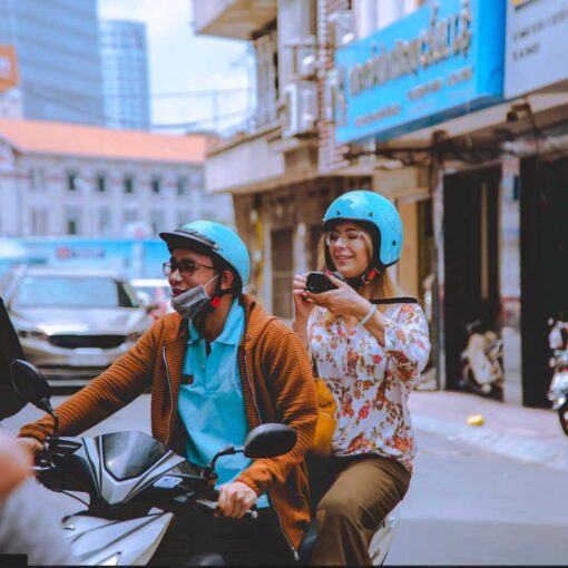A smiling tourist taking photos while riding on the back of a motorbike with a local student guide during a scooter tour in Ho Chi Minh City.