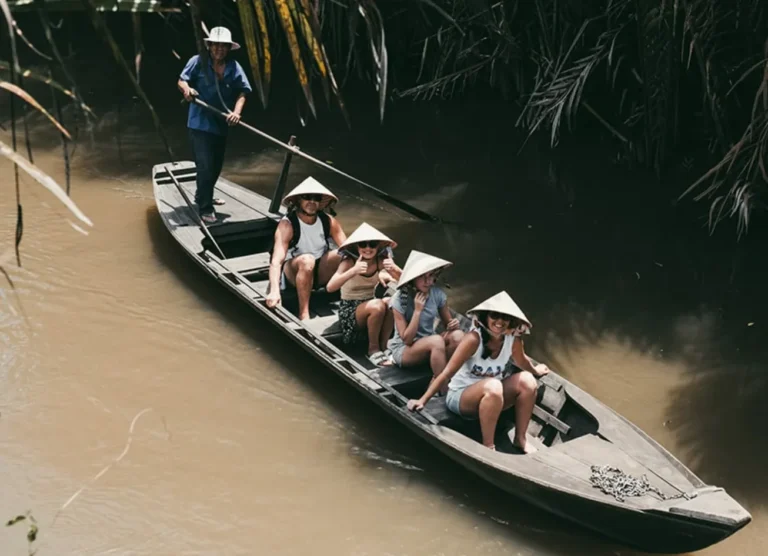 A group of travelers wearing traditional conical hats being rowed in a traditional boat along a lush, jungle-lined canal in the Mekong Delta, Vietnam.