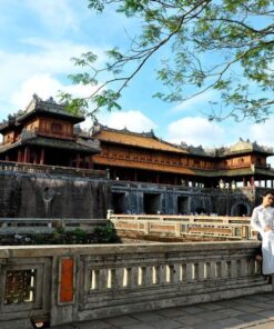 A woman in a traditional white Ao Dai sitting on the stone bridge of the Hue Imperial City (The Citadel) under a bright blue sky.