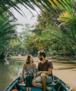 A smiling couple taking a scenic boat tour through a narrow, palm-fringed canal in the Mekong Delta, Vietnam.