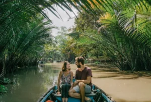 A smiling couple taking a scenic boat tour through a narrow, palm-fringed canal in the Mekong Delta, Vietnam.
