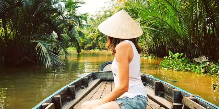 A female traveler wearing a traditional Vietnamese conical hat riding a wooden rowing sampan boat through a Mekong Delta canal.
