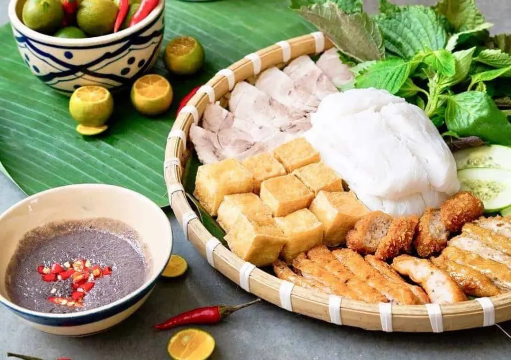 A traditional bamboo platter of Bún Đậu Mắm Tôm featuring rice vermicelli, crispy fried tofu, boiled pork, and fresh herbs, served with a bowl of pungent fermented shrimp paste (Mắm Tôm) and red chili. A highlight of the Wild and Weird Hanoi Street Food Tour.