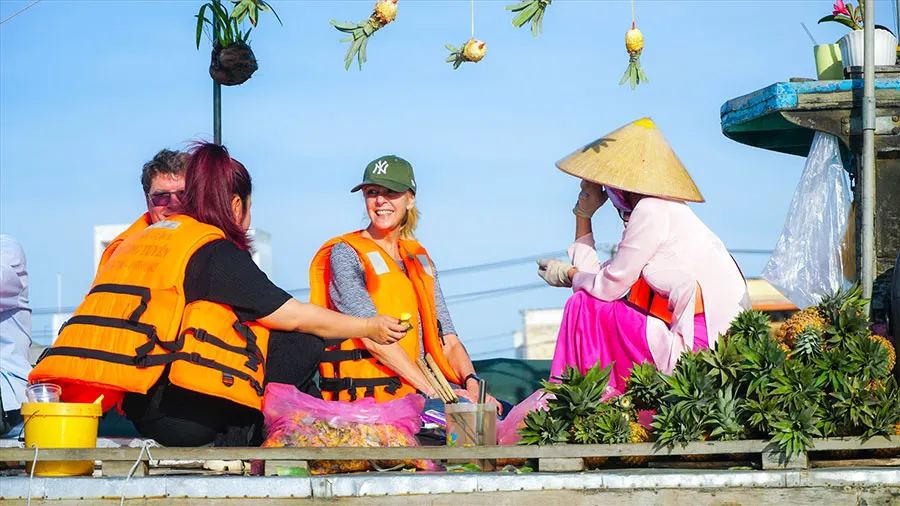 Foreign tourists experience eating pineapples on the boats of local vendors at Cai Rang floating market.